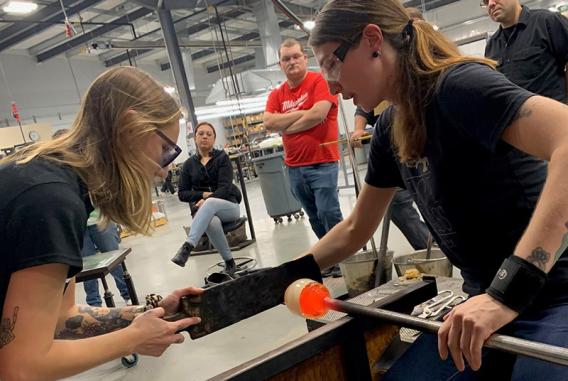 Two women working in a glassblowing studio with three others watching